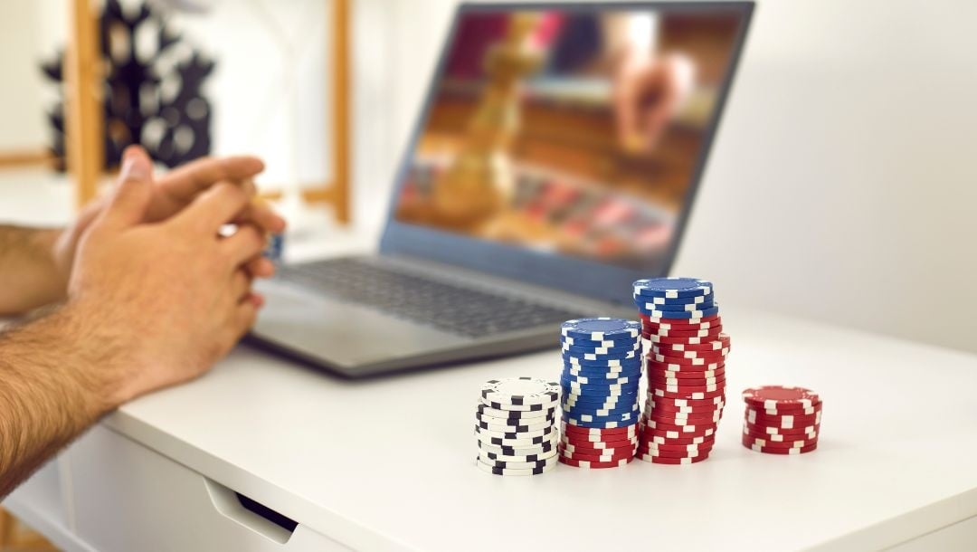 A person sitting at a desk, with red, blue, and white casino chips neatly stacked on top of the desk, next to a laptop with a game of roulette on the screen.