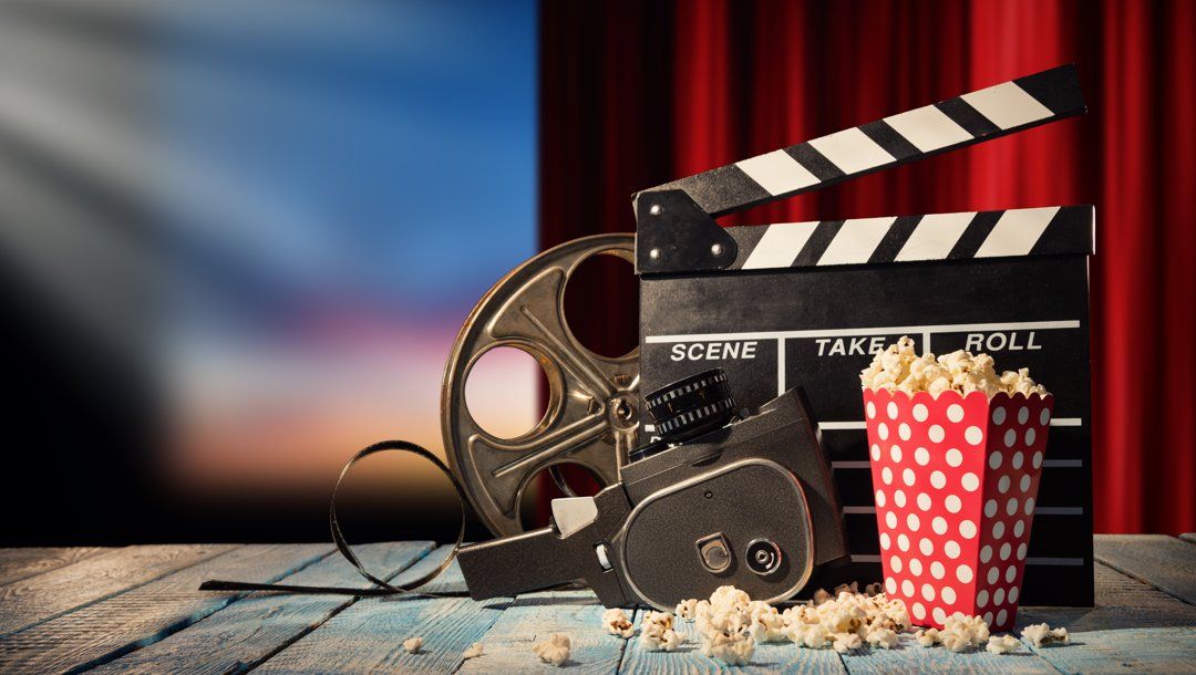 A clapper board, film reel, film camera, and box of popcorn in front of a red curtain and blurry cinema screen.