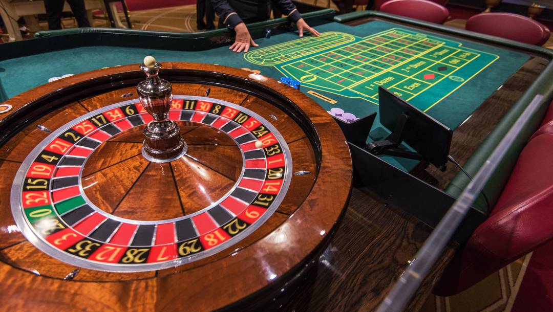 A casino dealer stands behind a green roulette betting table beside a polished wooden roulette wheel.
