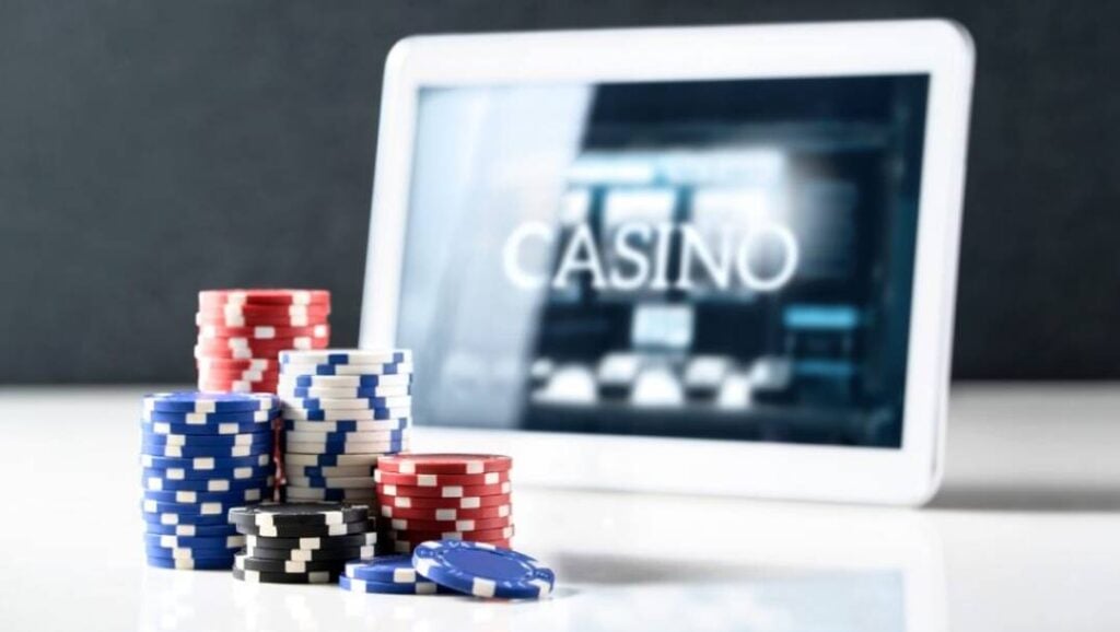 Stacks of colorful poker chips sit in the foreground next to a digital tablet displaying an online casino.