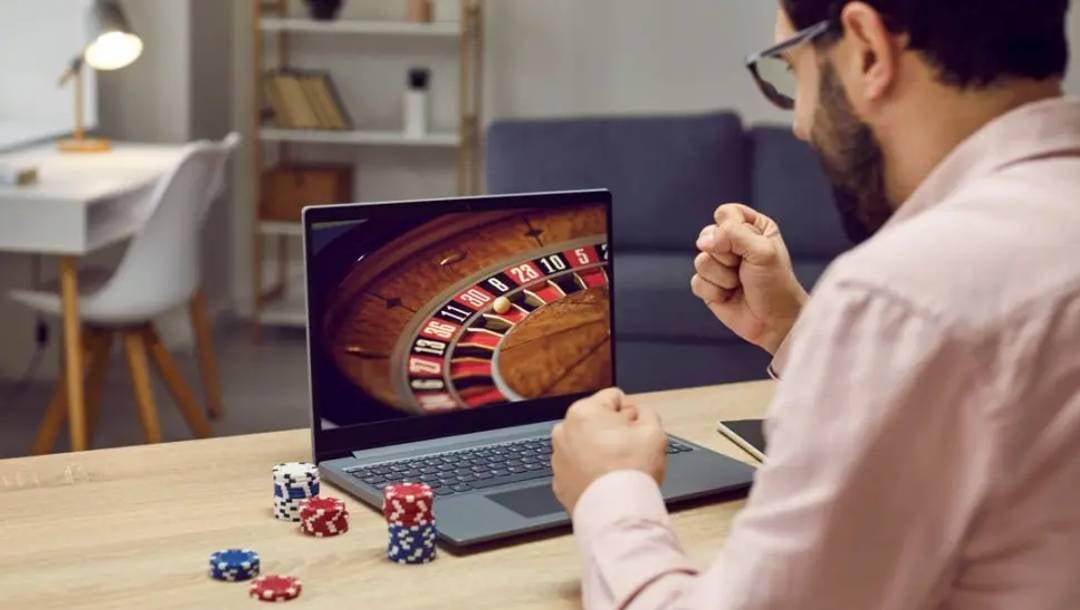 A man clenches his fists in celebration while playing an online roulette game on his laptop with casino chips stacked on his desk.