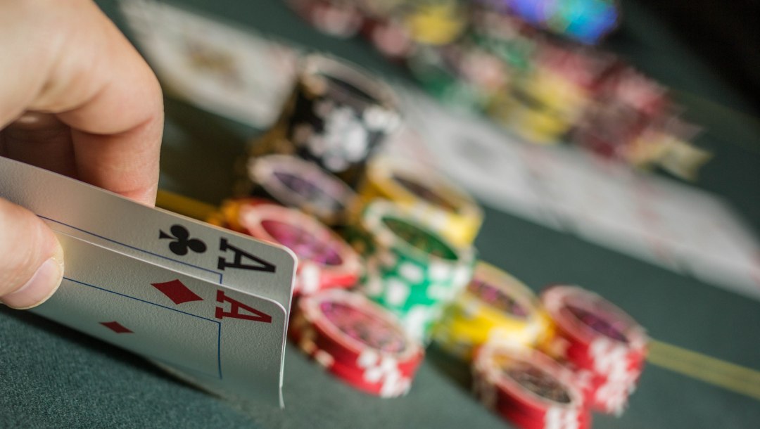 A hand holding two ace playing cards. There are casino chips and playing cards on the green felt table. The background is blurry.