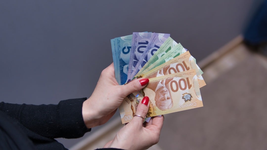 A woman is holding a stack of money in her hands. She has red nail polish on her nails. The background shows a wall and the floor.