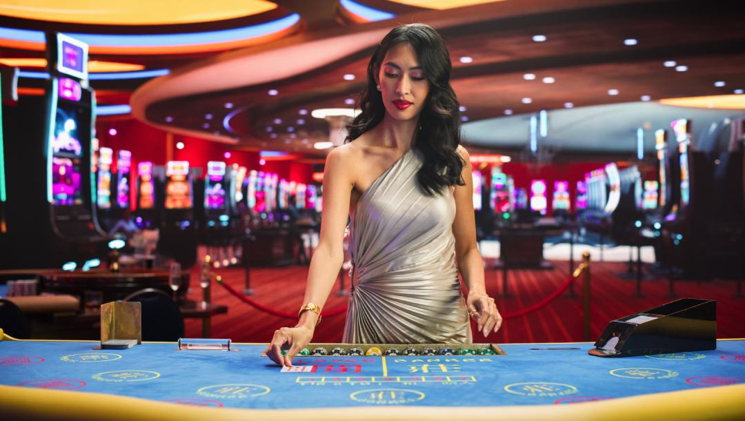 A woman in a red dress placing a playing card on a blue casino table. The background shows the inside of a casino floor with slot machines.