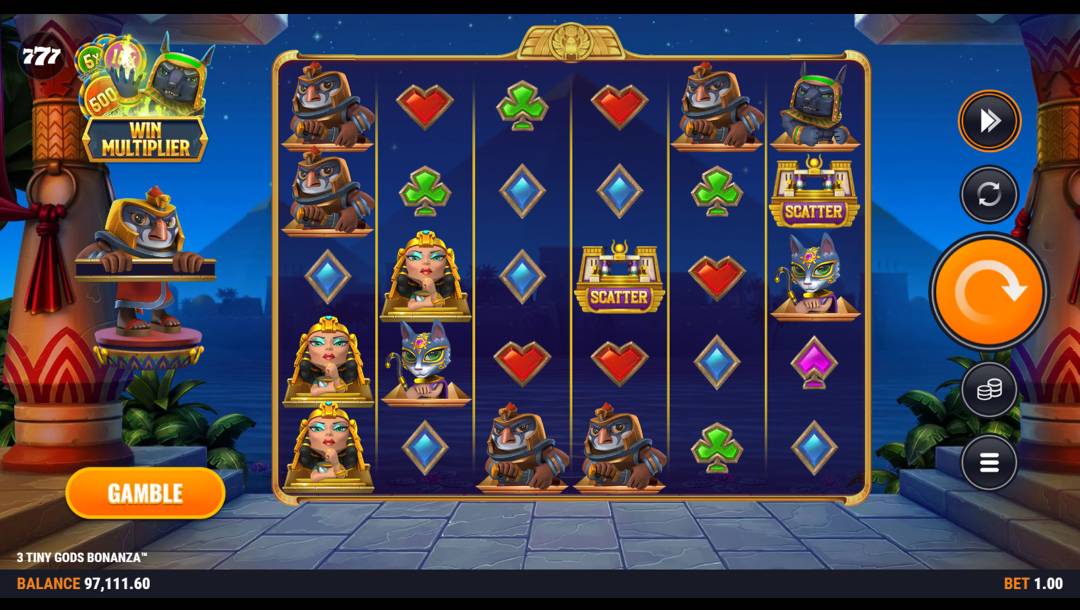 A screenshot of the slot reels in 3 Tiny Gods Bonanza. The backdrop is the entrance to an Egyptian building with the silhouette of an Egyptian city and pyramids visible against the night sky. In the foreground are the slot reels with a range of cute Egypt-themed symbols, including the Egyptian queen, Horus, Bastet, Anubis, and the Egyptian temple scatter. There are also regular symbols like the diamond, clubs, hearts, and spades symbols. To the left of the reels, Horus waits to unfurl his parchment.