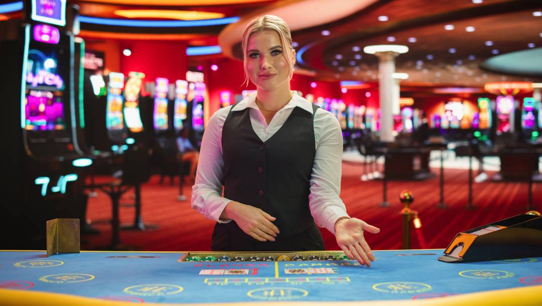 A female dealer in a black waistcoat, white collared shirt, and black slacks gestures to a player to sit at a casino card table. There are dozens of slot machines and other casino game tables behind her.