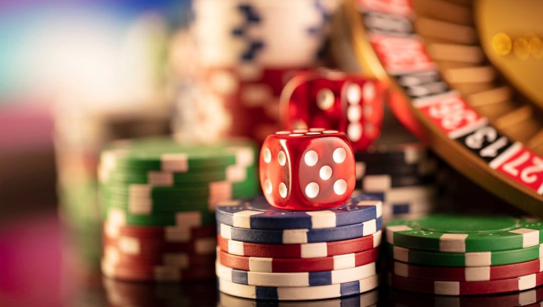 A close-up of a pair of dice on a stack of casino chips next to a small roulette wheel. There are other stacks of casino chips around the dice and roulette wheel.