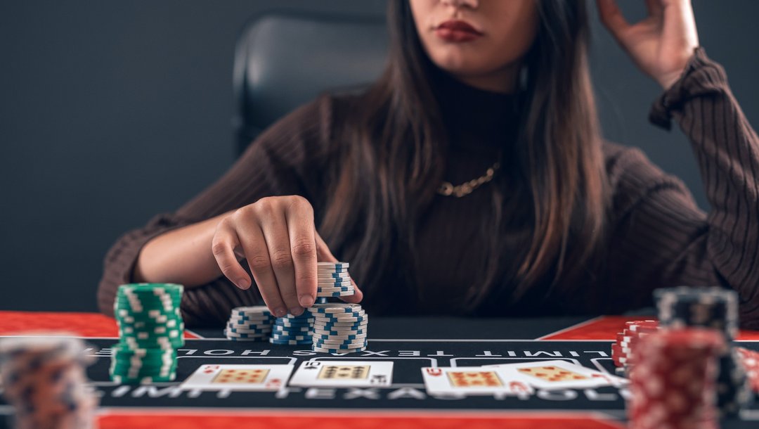 A woman is seated at a poker table, placing a bet with a stack of white and blue casino chips in the foreground. She is wearing a dark brown, ribbed turtleneck sweater and a gold necklace, with her face partially out of frame to the right. The playing cards lay face-up, with various chip stacks in green, black, and red colours around them. In the background, a dark, minimal setting with a black chair.