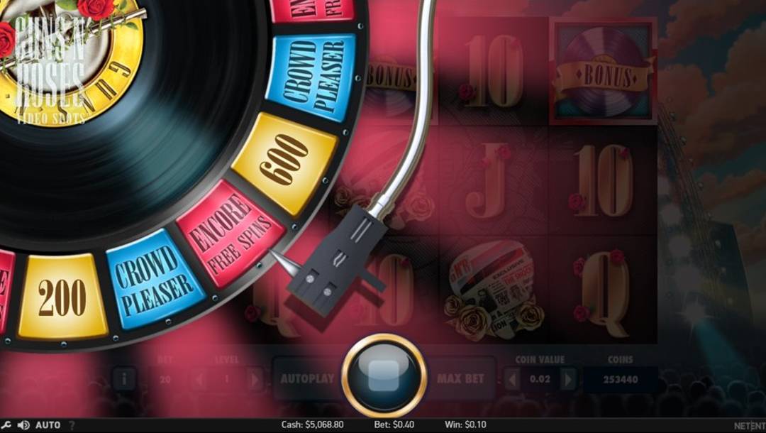 A screenshot of the Bonus Wheel feature from the Guns N’ Roses online slot. It features a large spinning vinyl record wheel on the left side labeled with rewards like Crowd Pleaser, Encore Free Spins, and coin values like 200 and 600. A stylized record-player arm points to a winning segment while the background shows faded slot reels and band-themed symbols, including roses and golden letters. The stage lighting and concert crowd beneath a pink spotlight haze evoke a rock concert atmosphere, with a performance stage and towering speakers visible in the backdrop.