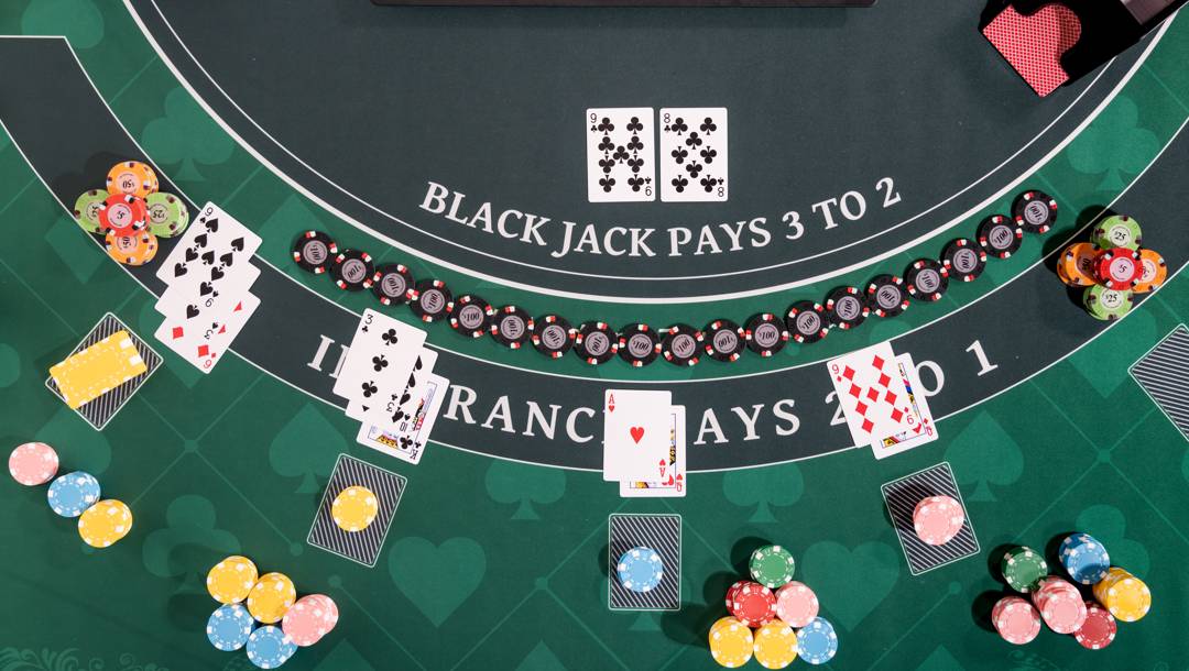 A top-down view of a blackjack table showing several player hands with face-up cards and colorful stacks of chips placed next to each hand. The dealer’s hand is visible at the center, as are four other players’ hands at various positions around the table. The player’s bets are on the table behind their hands.