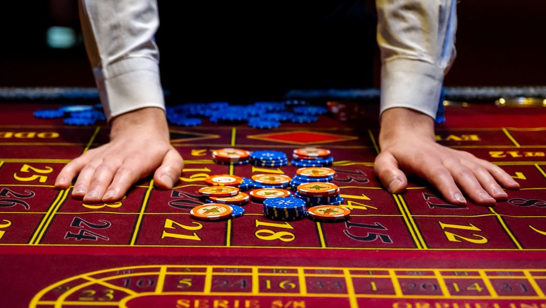 A casino dealer in a white dress shirt plants both hands on a burgundy roulette layout in the foreground, guarding neat stacks of multicoloured chips piled across yellow-outlined numbers 17, 20, and 25. Behind the croupier’s arms, the background fades into soft focus, allowing the vivid felt and chips in the foreground to command attention.
