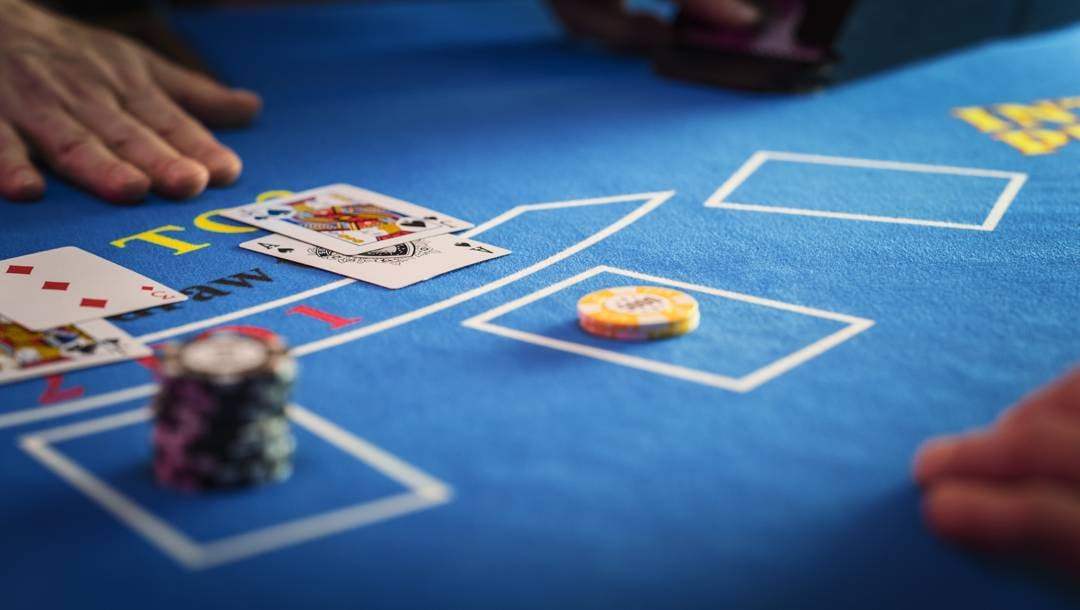 A close-up of a blue baccarat table with playing cards, casino chips, and players’ hands around the betting area.