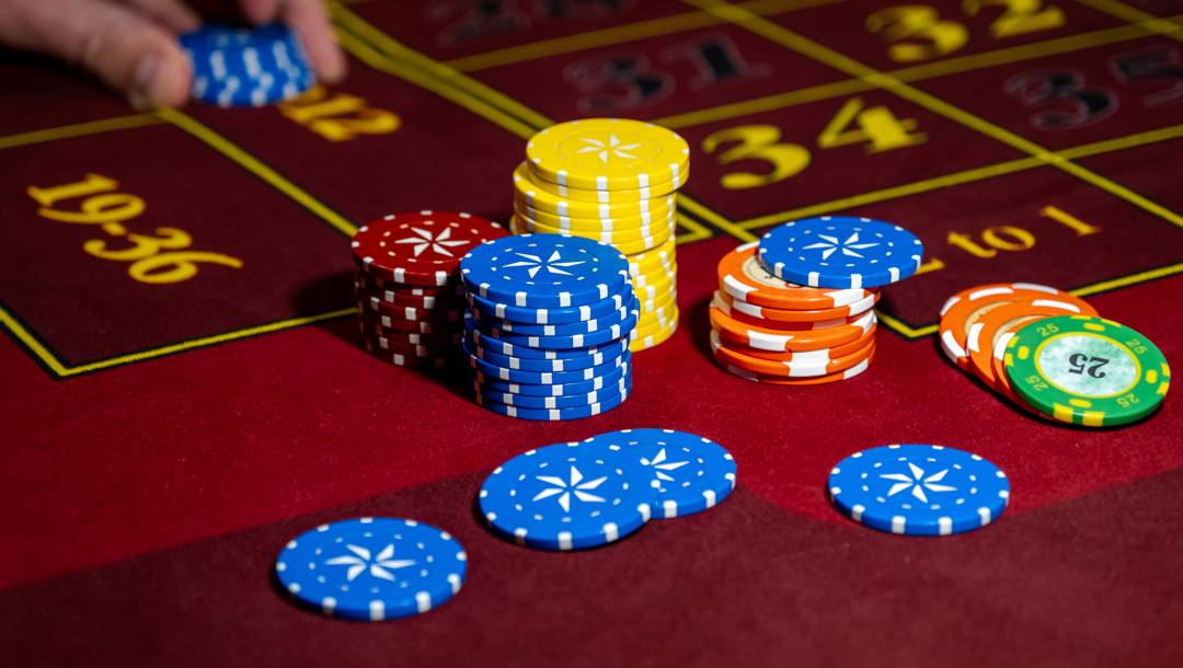 Stacks and scattered multicoloured casino chips rest on a red roulette betting table as a player places a wager.
