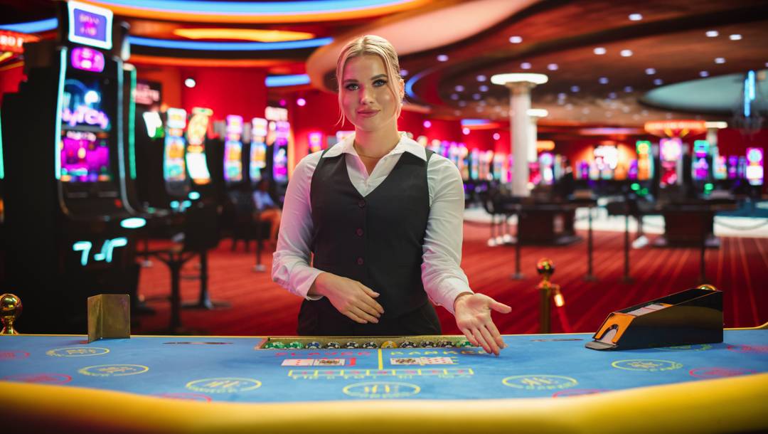 A female casino dealer gestures toward a baccarat table in a brightly lit casino with slot machines in the background.