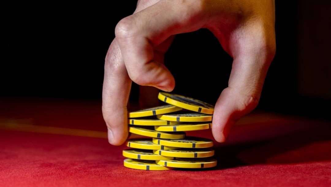 A hand riffles a stack of yellow casino chips on a red felt table against a dark background.