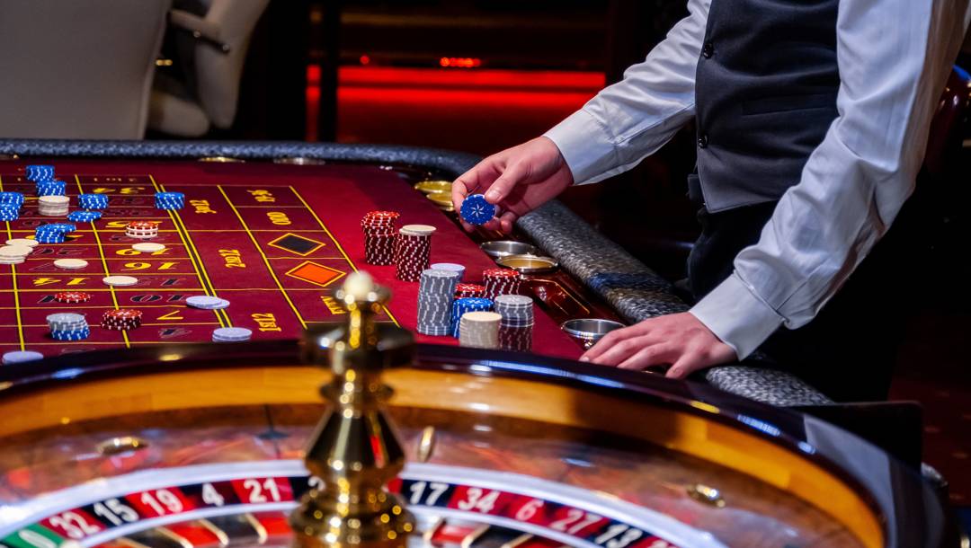 A roulette dealer arranges stacks of colourful chips beside a roulette wheel on a red casino table.